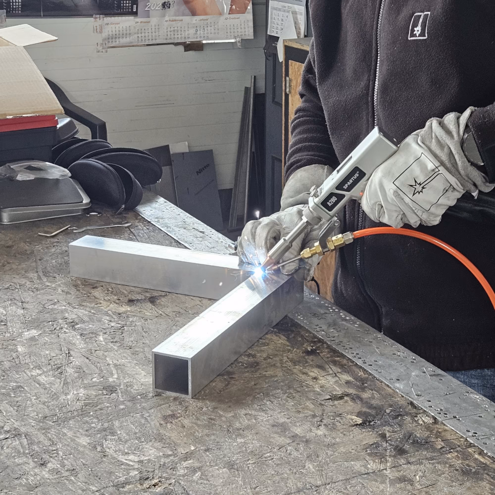 Spawacz w rękawicach ochronnych używający ręcznej głowicy spawarki laserowej SPARTUS do precyzyjnego łączenia metalowych profili na stole warsztatowym.